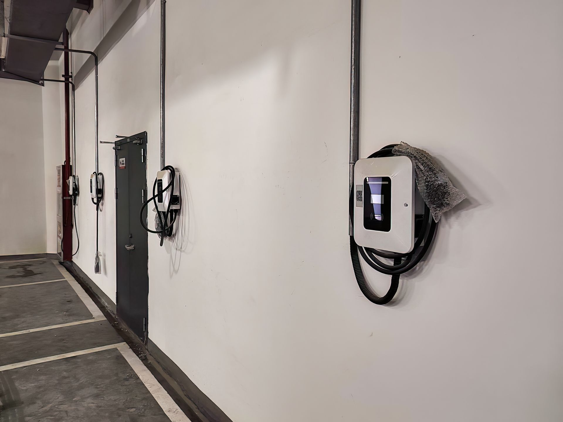 Electric vehicle chargers mounted on a white wall in a parking garage. Cables and conduit are visible.