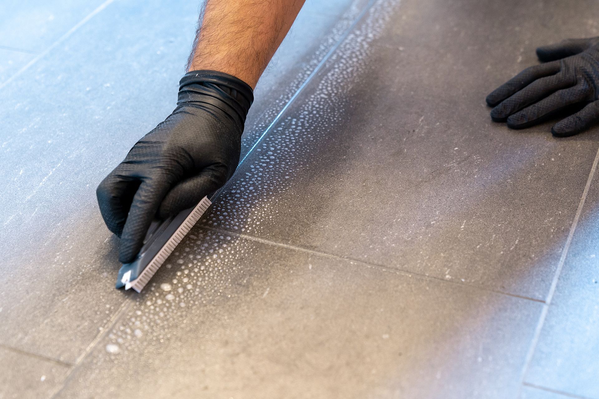 Person wearing black gloves cleaning a tiled floor with a squeegee.