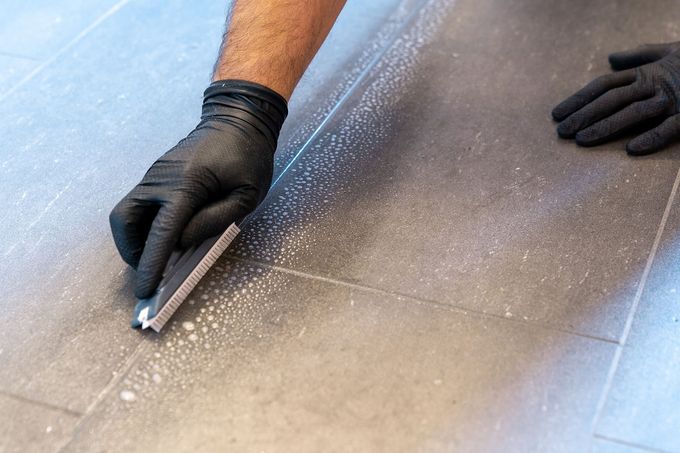 Person wearing black gloves cleaning a tiled floor with a squeegee.