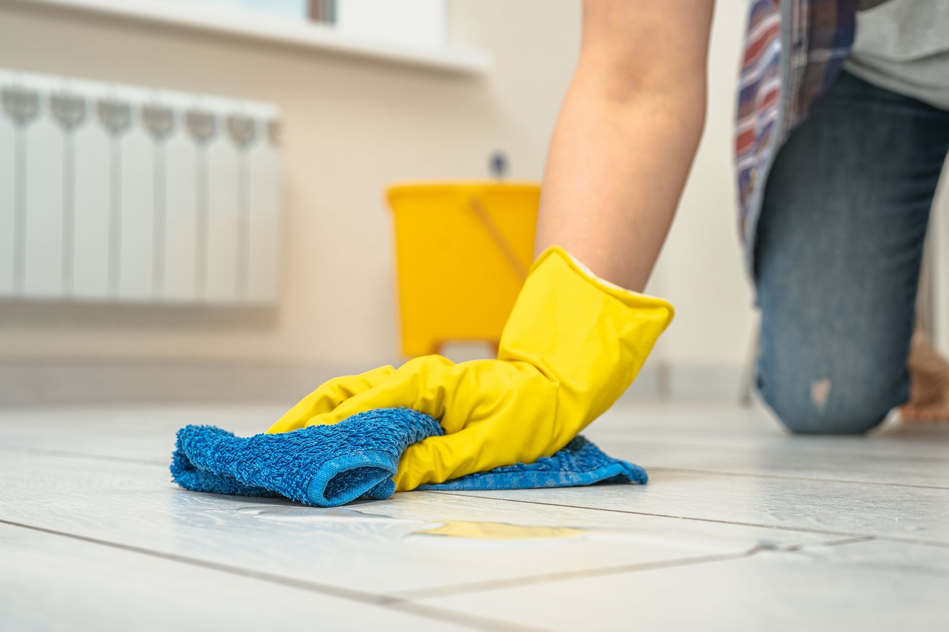 Person wearing yellow gloves wiping a tiled floor with a blue cloth, yellow bucket in the background.