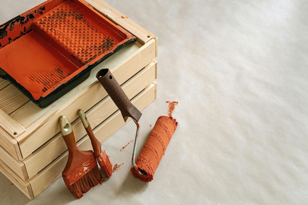 Paint tray, brushes, and roller with orange paint on wooden crate and neutral surface.