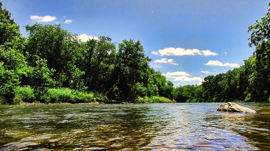 River flowing between green trees under a blue sky with clouds.