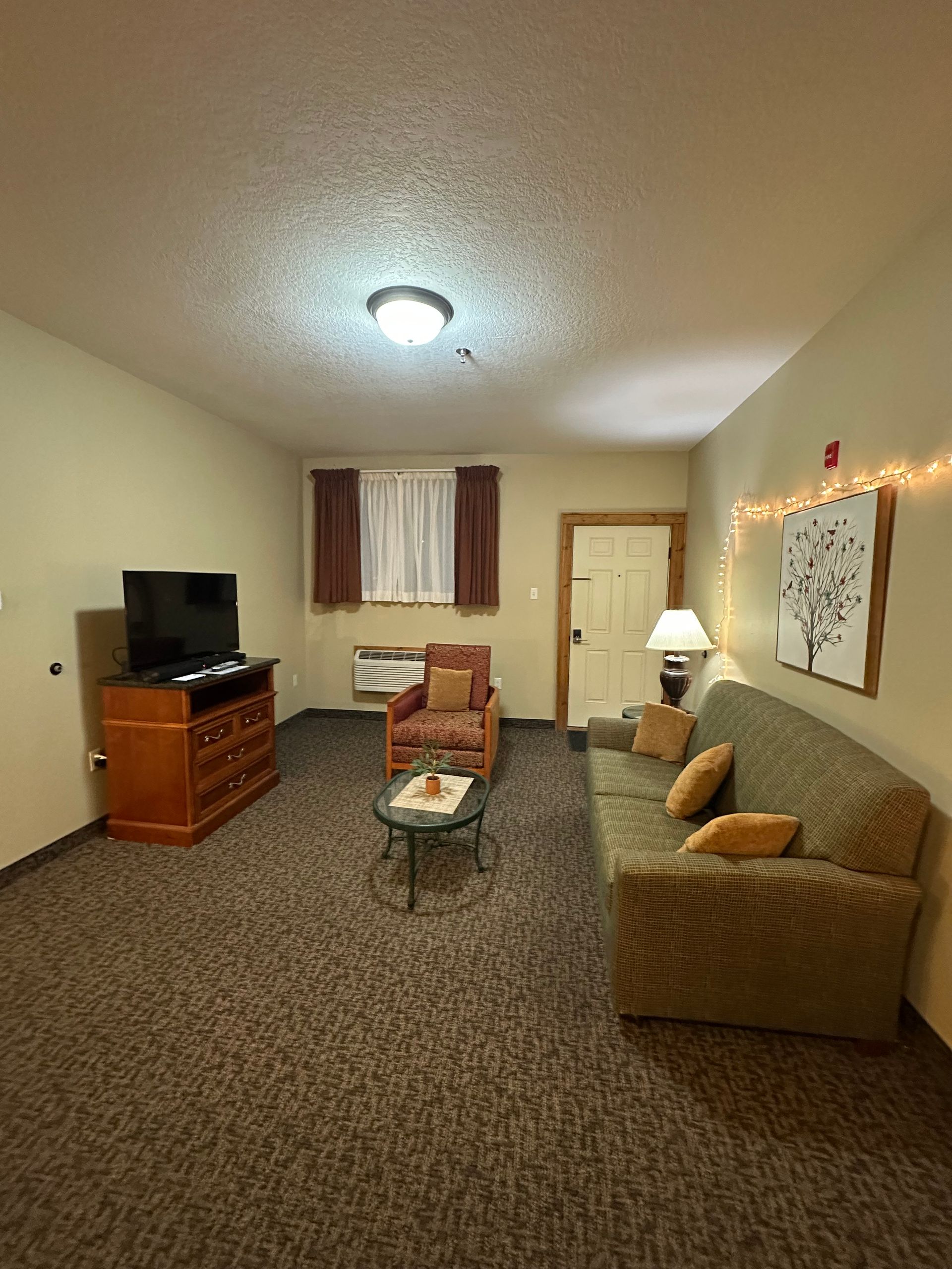Living room with TV, sofa, armchair, coffee table, and doorway. Beige walls and carpet.