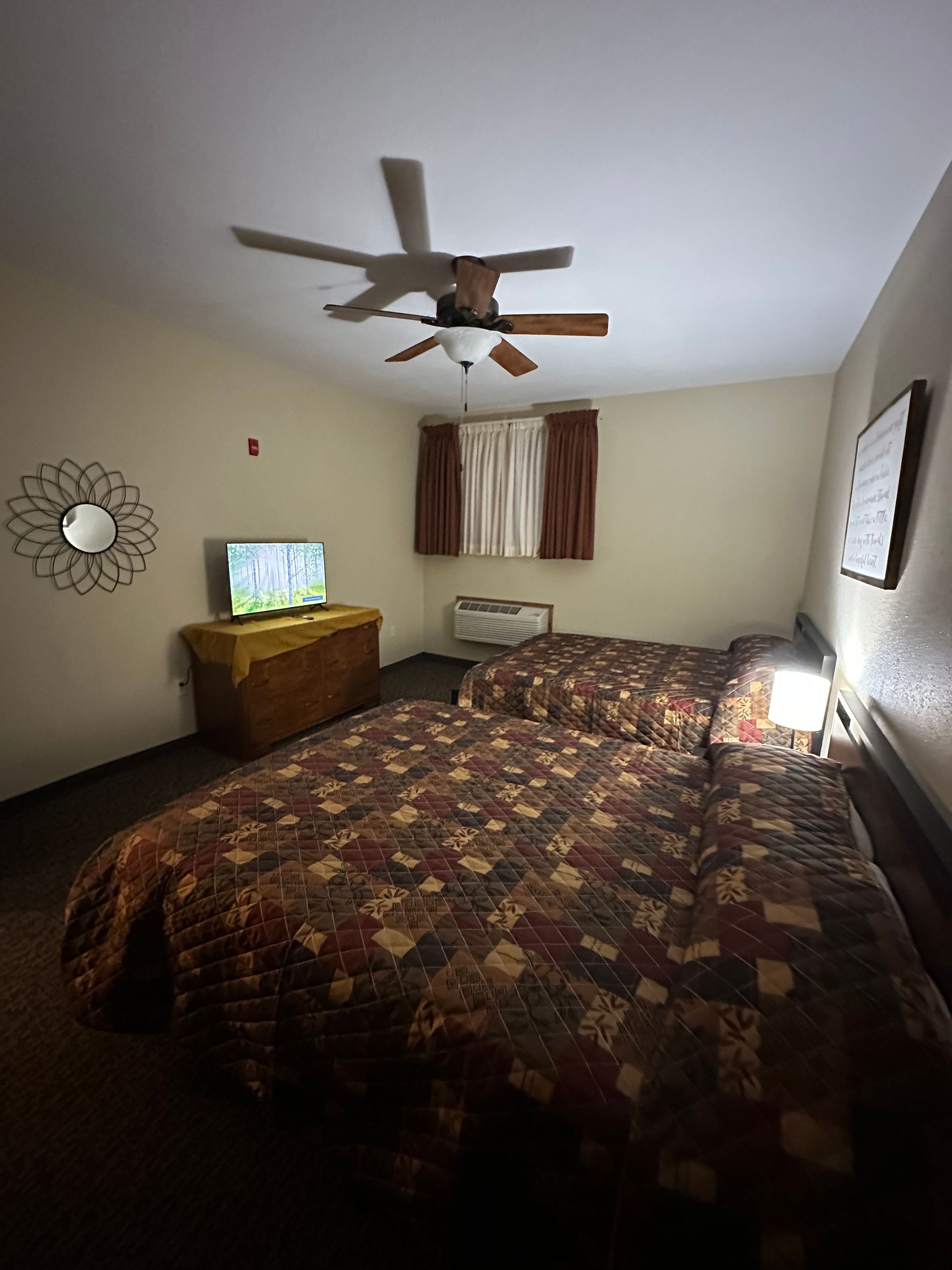 A motel room with two beds, a TV, mirror, and ceiling fan. Dark brown and red bedding. Beige walls.