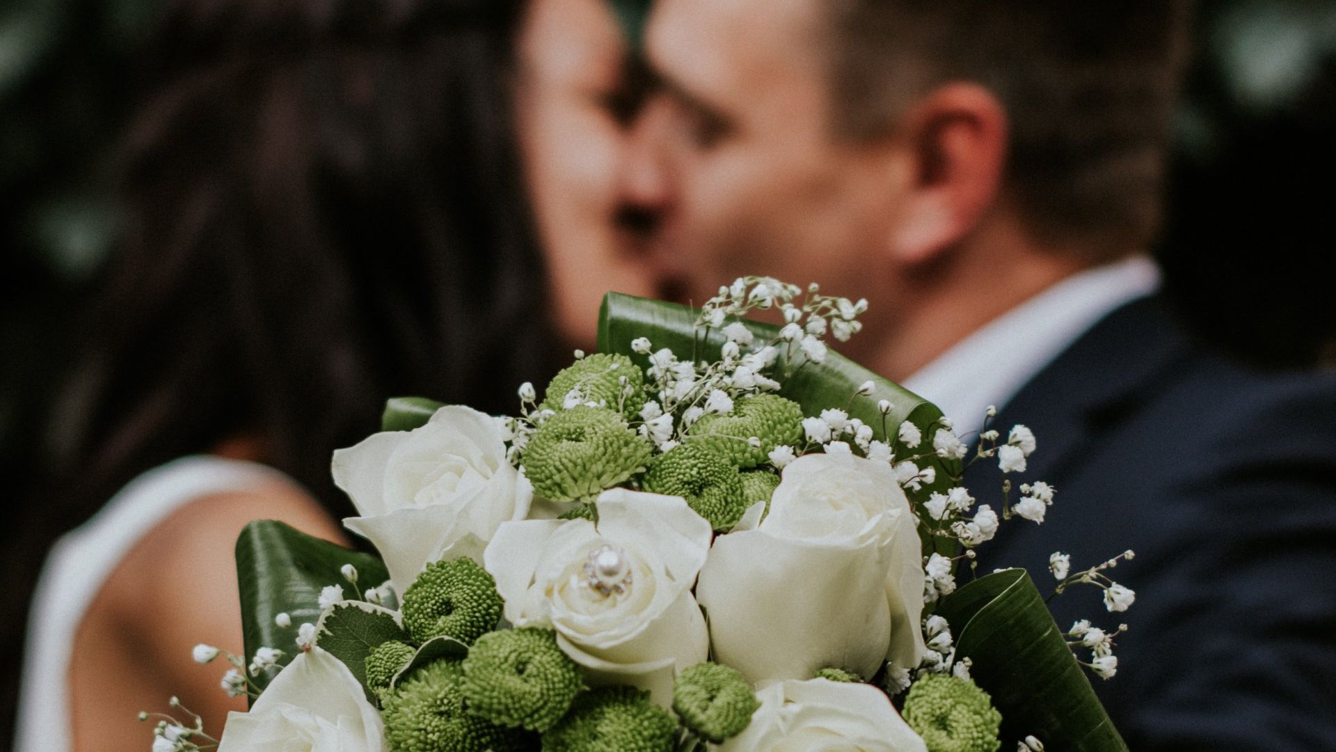 Wedding bouquet in focus; blurred couple kissing in background. White roses, green accents.