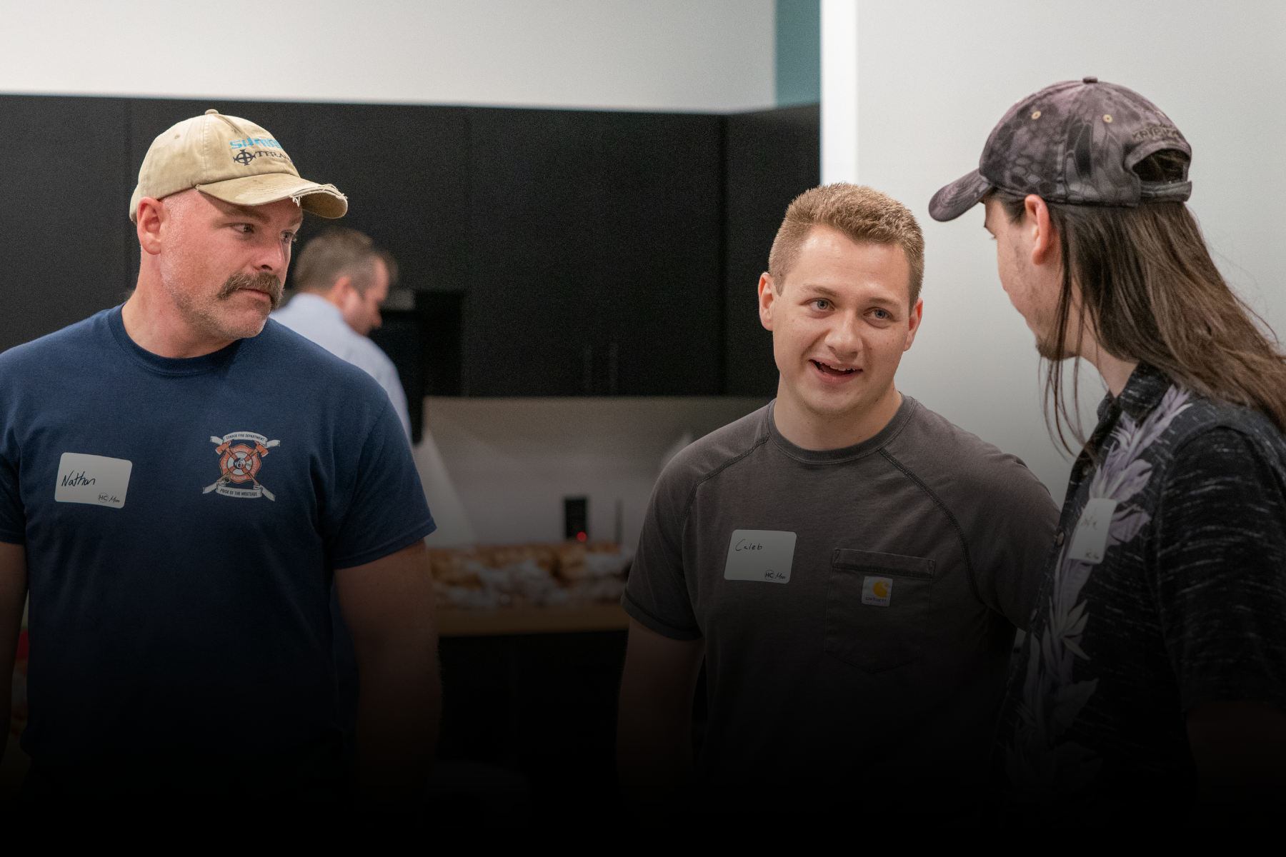 Three people talking indoors, two facing a smiling man in a black shirt and cap.