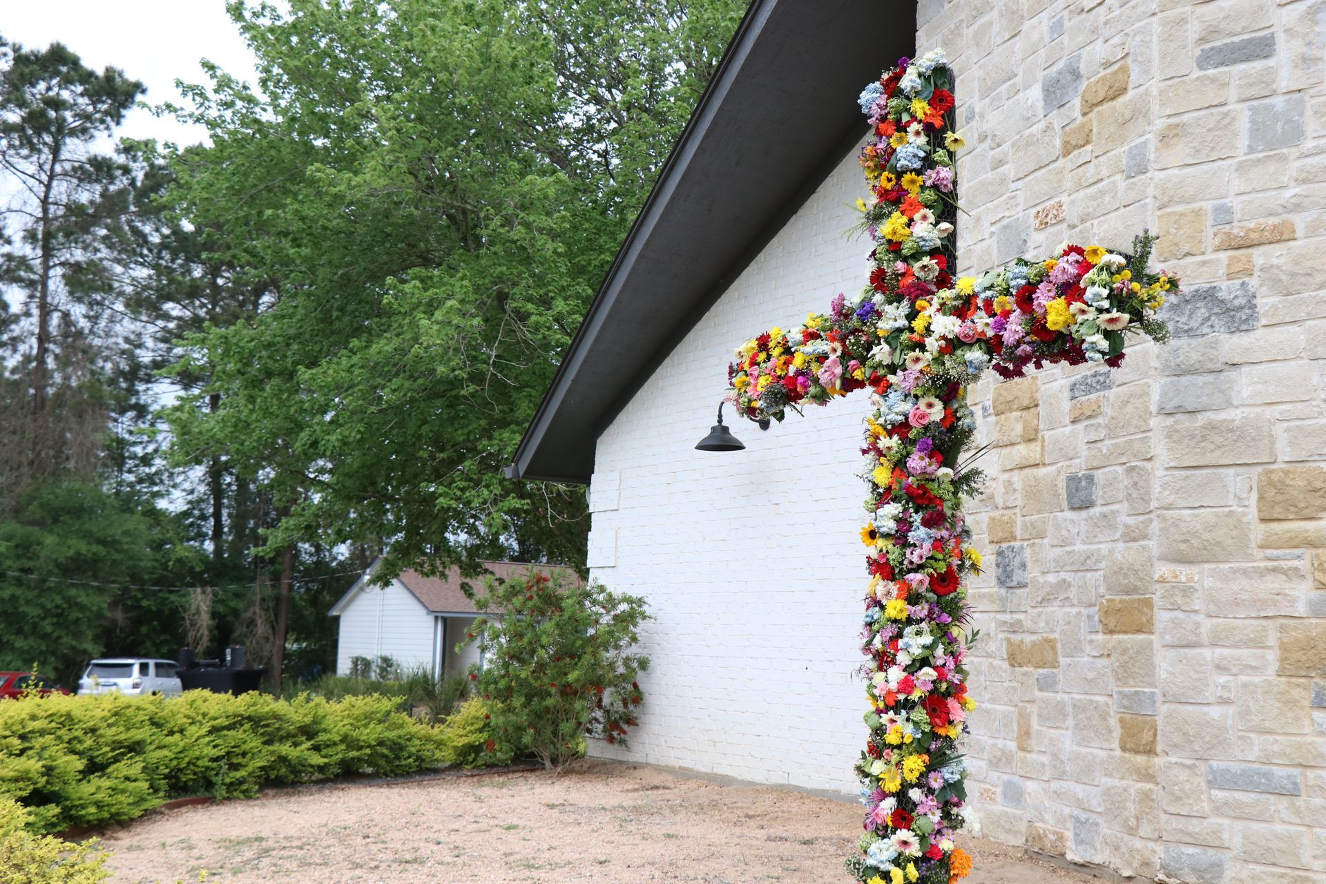 Flower-covered cross on a stone church wall beside a gravel path and trees