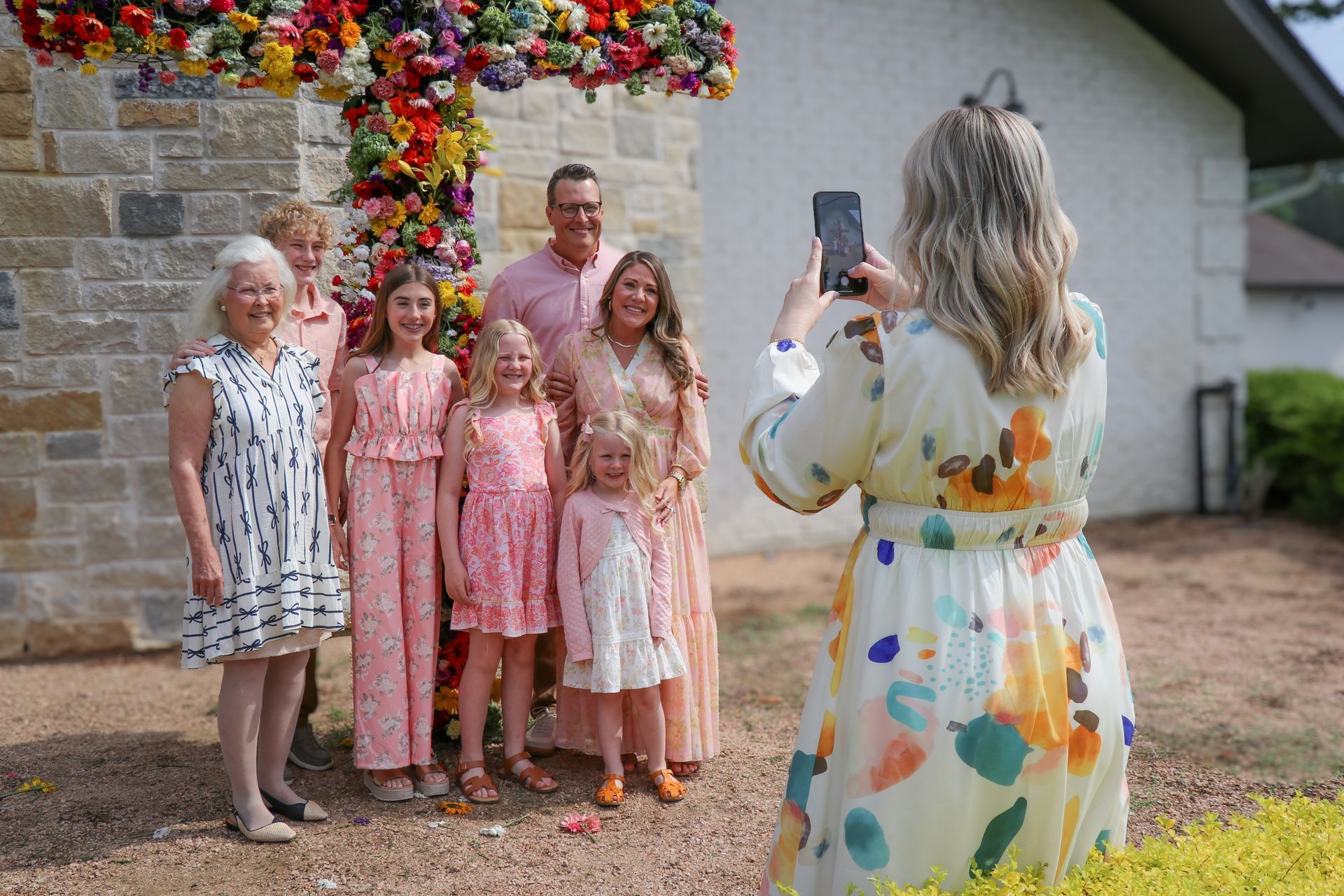 Woman in floral dress taking a photo of four people posing outdoors by a flower-covered arch.