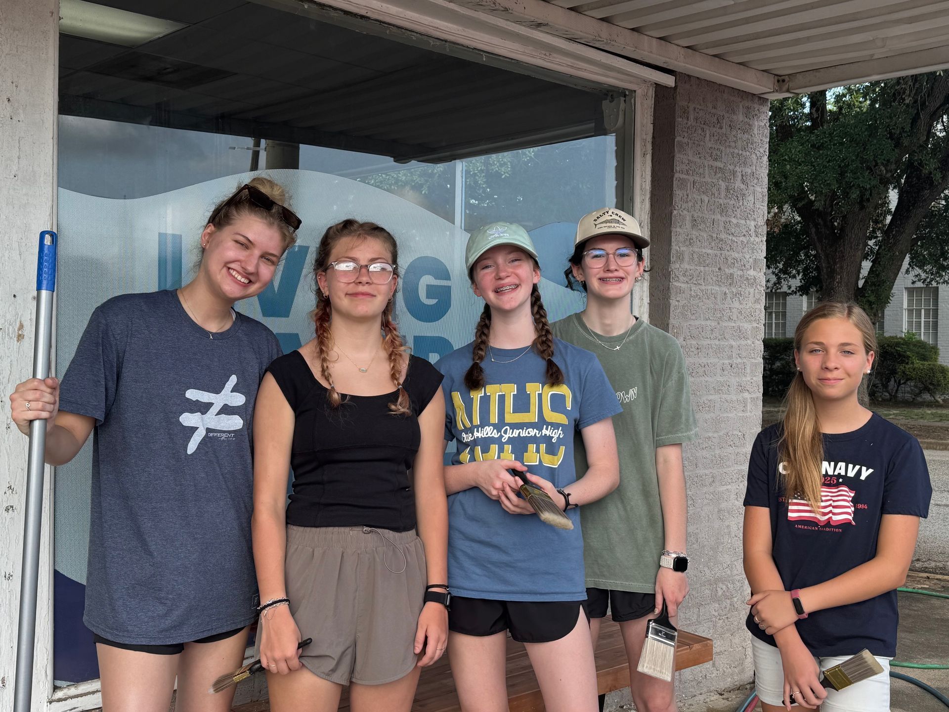 Five people standing and smiling under a porch, holding a broom and a propeller hat.