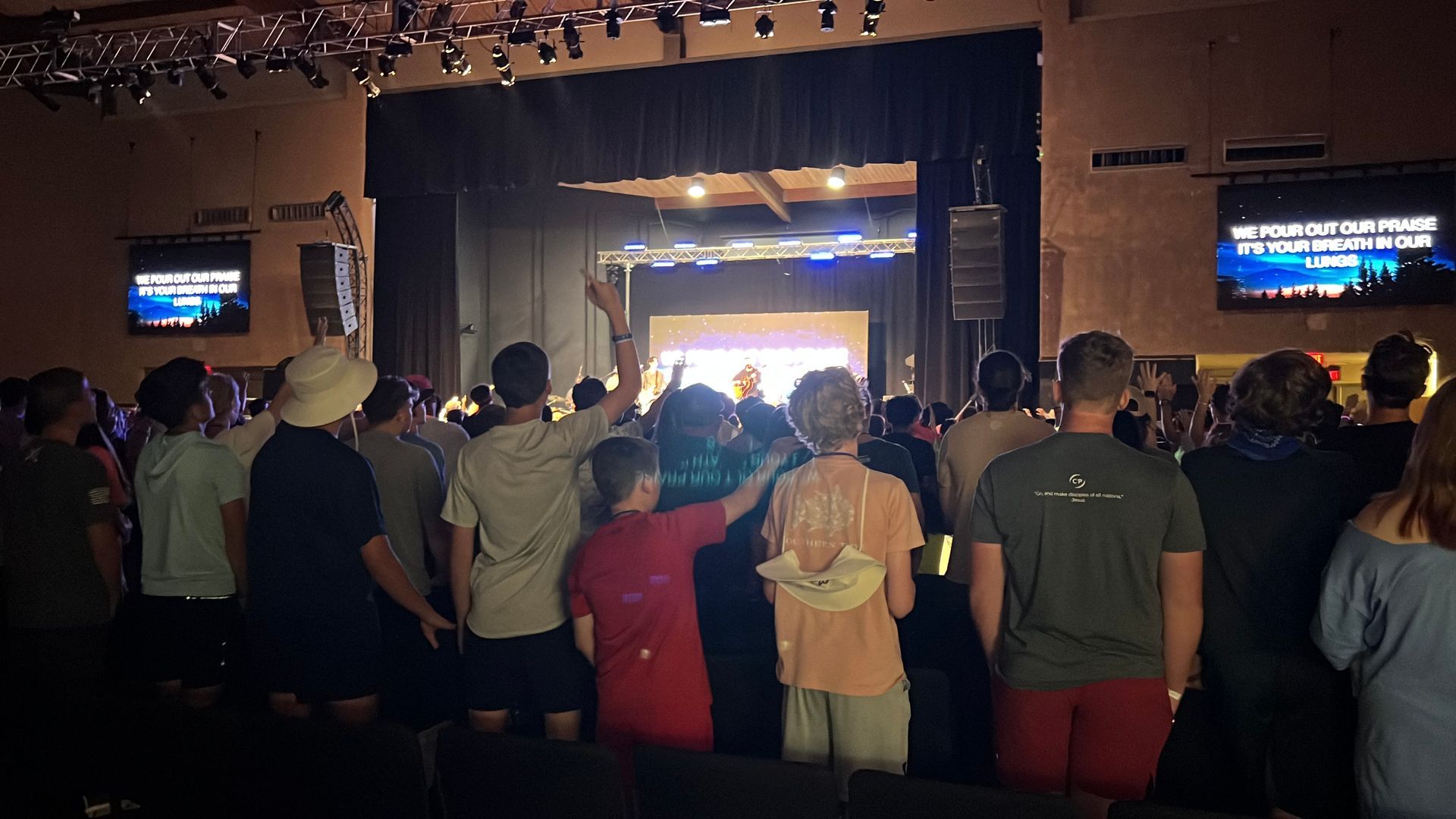 Crowd standing in a dim venue facing a brightly lit stage with large screens on both sides