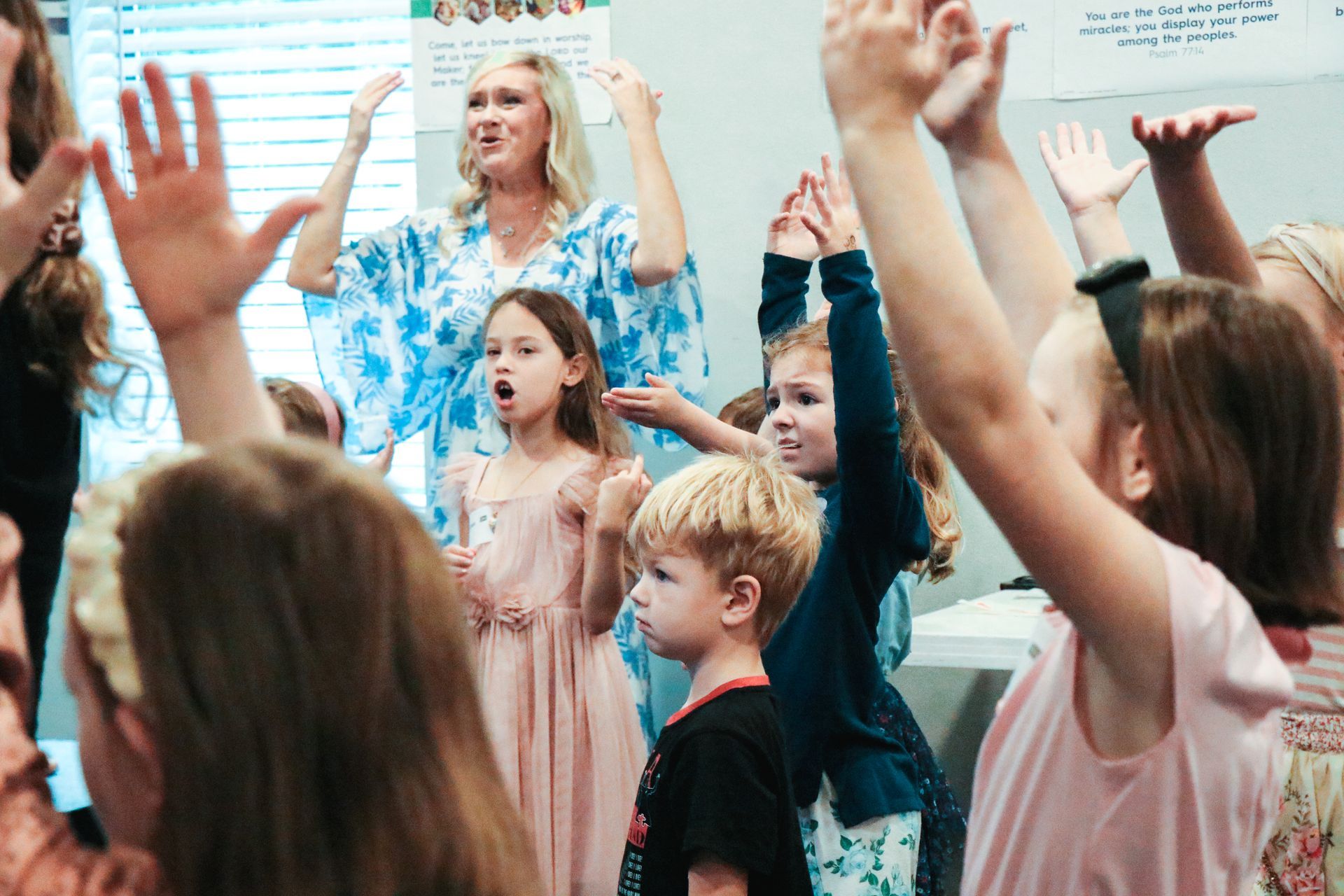 Children with raised hands in a classroom, led by an adult in a blue patterned shirt