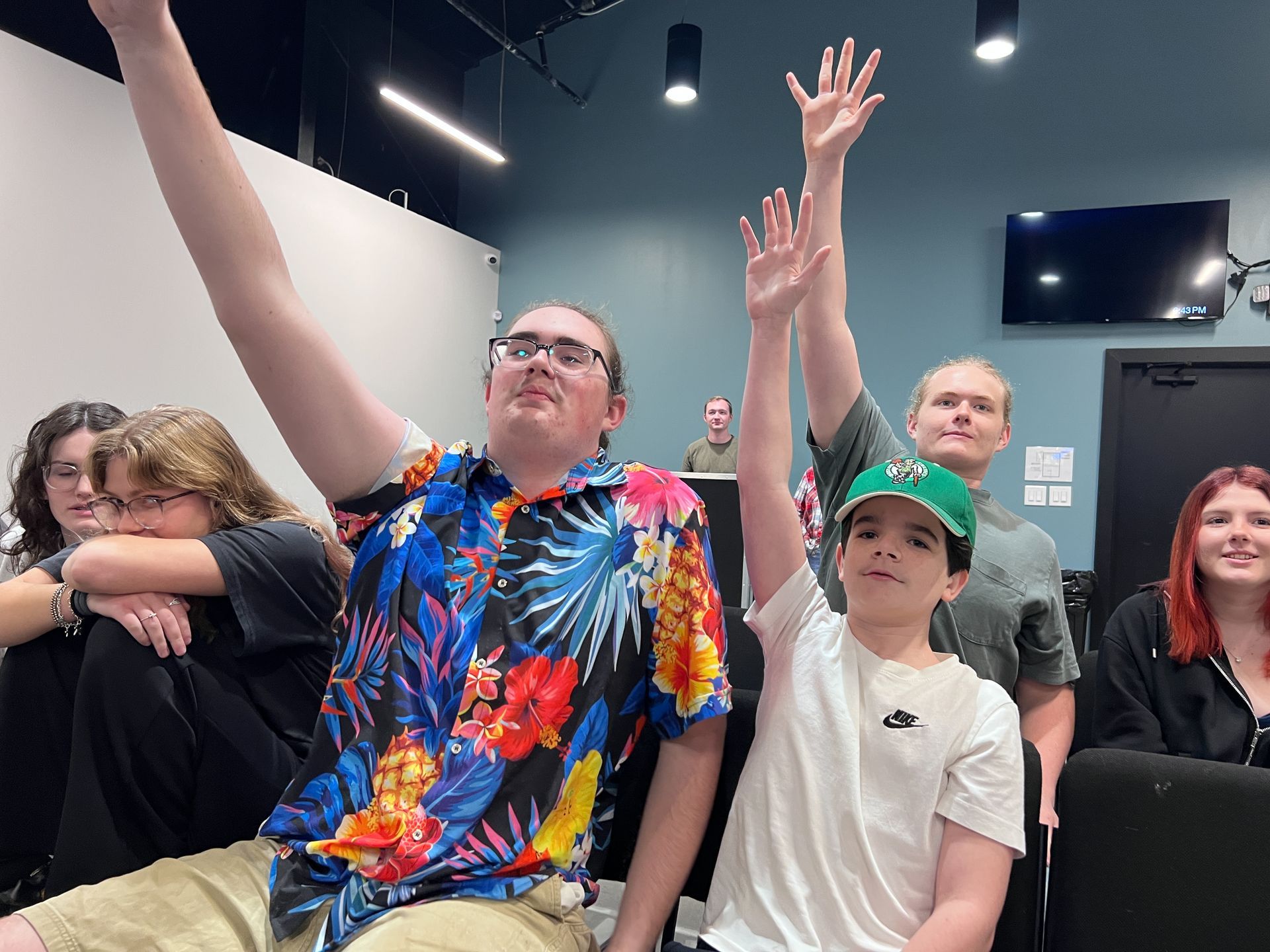 People seated in a room raise their hands, with colorful shirts and a black ceiling with spotlights above.