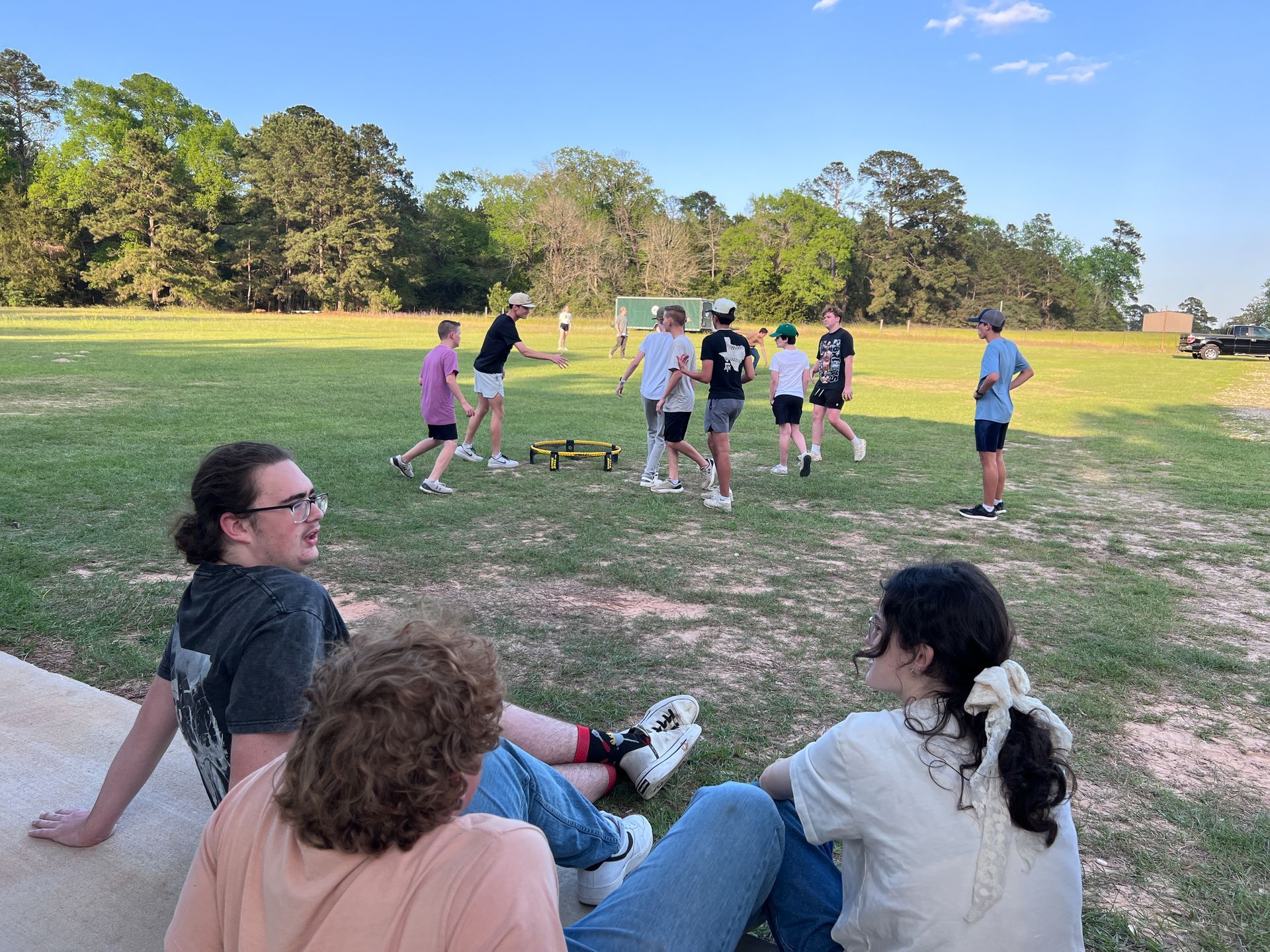 People seated near a grassy field watching a group exercise or game outdoors under a blue sky.