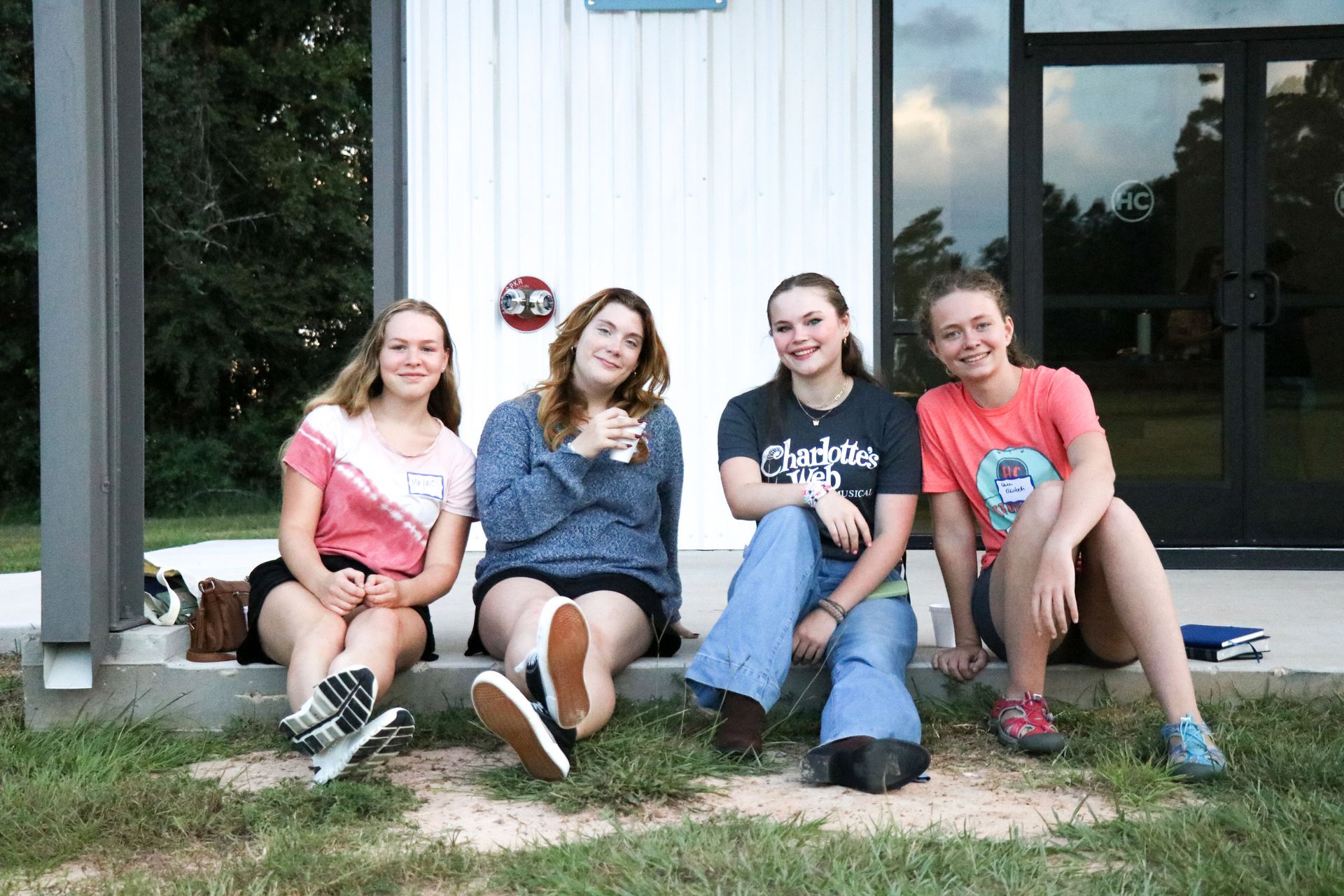 Four people sitting on grass in front of a white building, smiling at the camera.