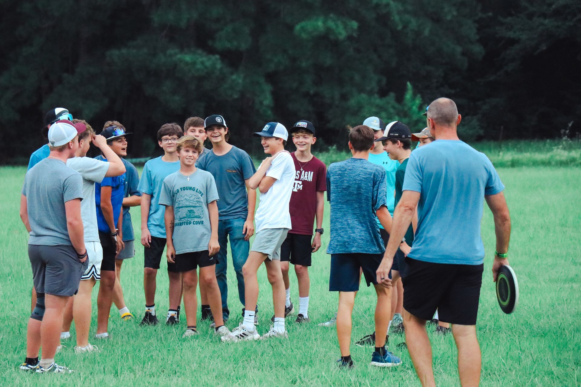 Group of people in athletic clothes gathered on a grassy field with a coach, likely for a sports practice.