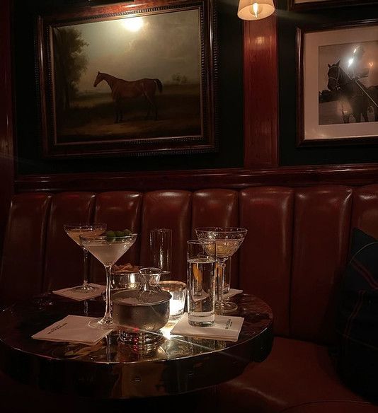 A small, candlelit round table with cocktails and glasses sits in a dimly lit, upscale lounge with leather booths.