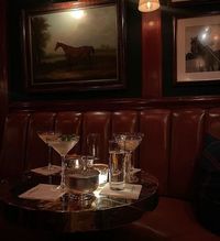 A small, candlelit round table with cocktails and glasses sits in a dimly lit, upscale lounge with leather booths.