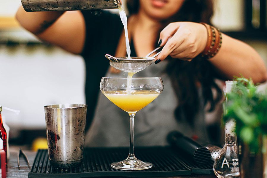 A bartender uses a fine mesh strainer to pour a yellow cocktail into a glass.