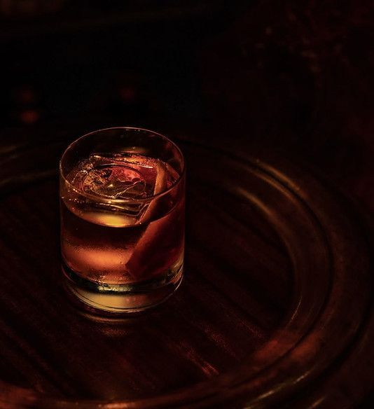 A rocks glass containing a dark amber cocktail with a large ice cube and citrus peel, centered on a dark wooden surface.
