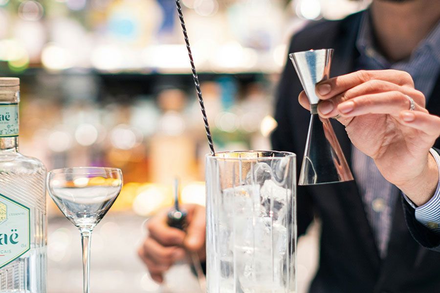 A person in a suit prepares a cocktail, holding a jigger over a glass of ice at a bar.