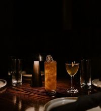 Two cocktails, one in a tall glass and one in a coupe, sit on a dark wooden table beside a small, glowing table lamp.