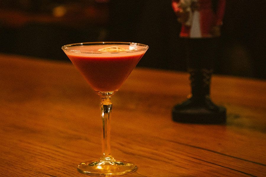 A pink cocktail in a stemmed martini glass sits on a wooden bar next to a blurred figure of a soldier figurine.