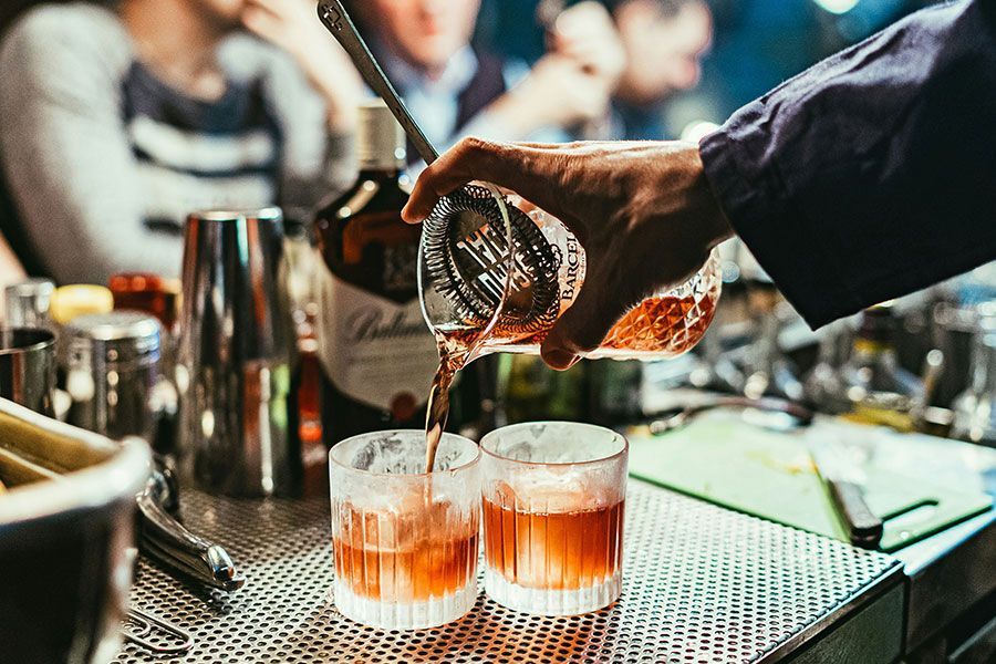 A bartender pours an amber cocktail from a mixing glass into two rocks glasses filled with ice on a metal bar mat.