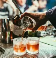 A bartender pours an amber cocktail from a mixing glass into two rocks glasses filled with ice on a metal bar mat.