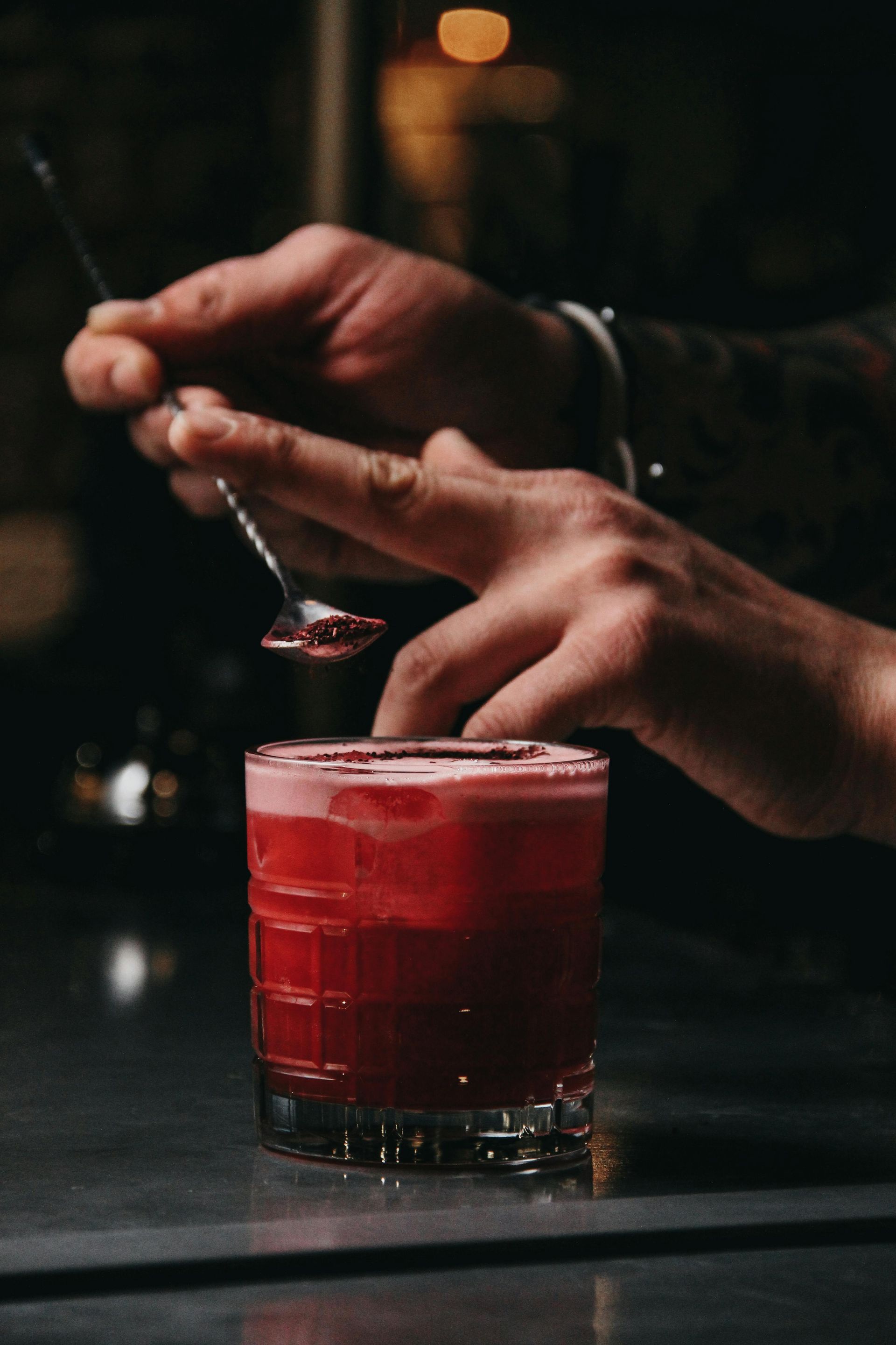 A bartender holds a long spoon over a vibrant red cocktail in a textured glass on a dimly lit bar counter.