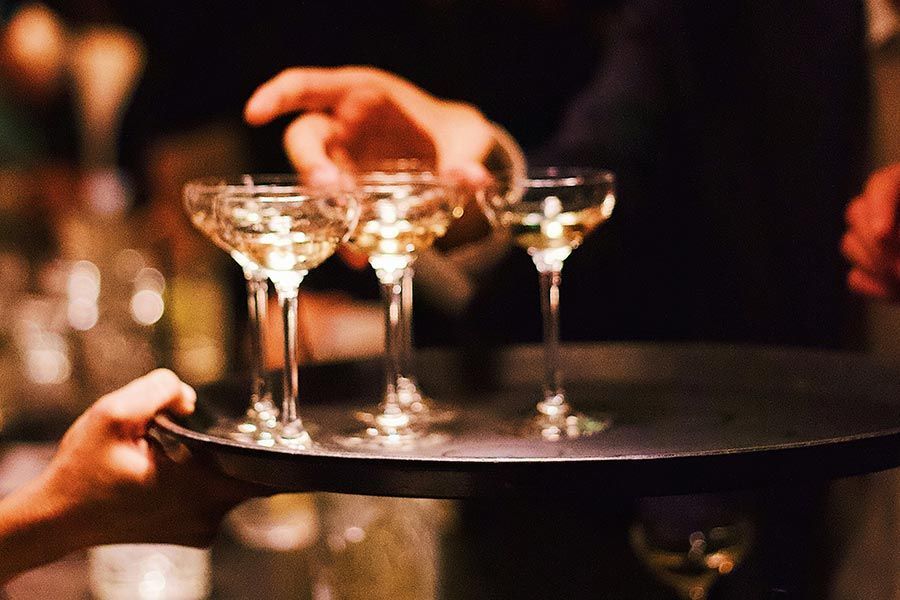A server holds a dark tray with several filled champagne coupes as a person reaches for a glass in a dimly lit setting.