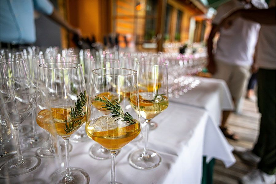 Rows of wine glasses filled with white wine and a sprig of rosemary arranged on a white tablecloth at an outdoor event.
