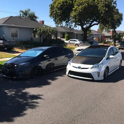 Two modified cars parked on a street, one black, one white with a carbon fiber hood, under a sunny sky.