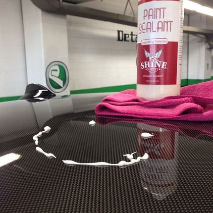 Paint sealant bottle on carbon fiber car panel, white liquid spilled, pink cloth, shop background.