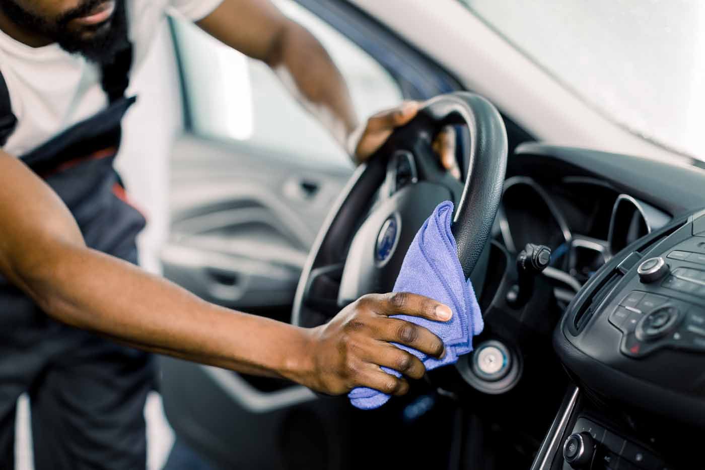 A man cleaning a car's black steering wheel with a blue cloth inside the vehicle.