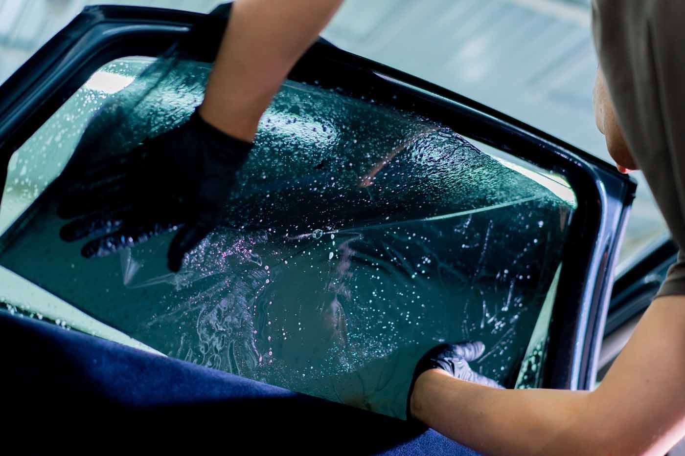 Hands applying window tint film to a car window. Black gloves, tint, and vehicle interior.