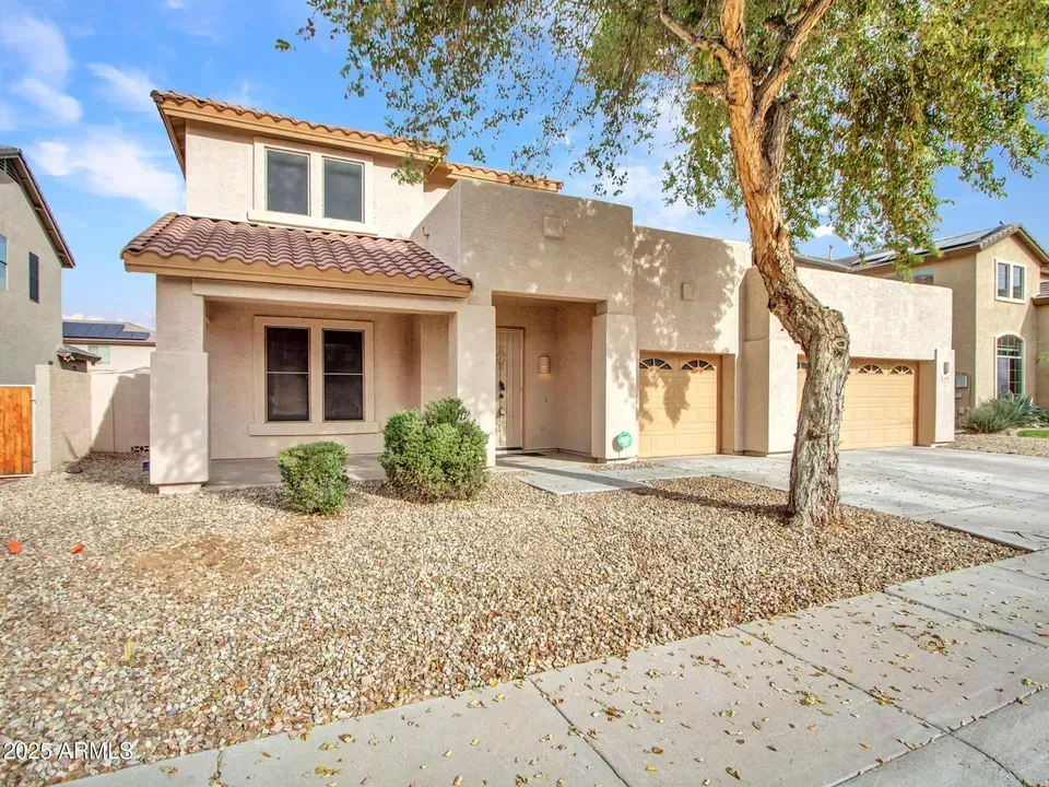 Tan stucco house with three-car garage, gravel yard, and tree in front.