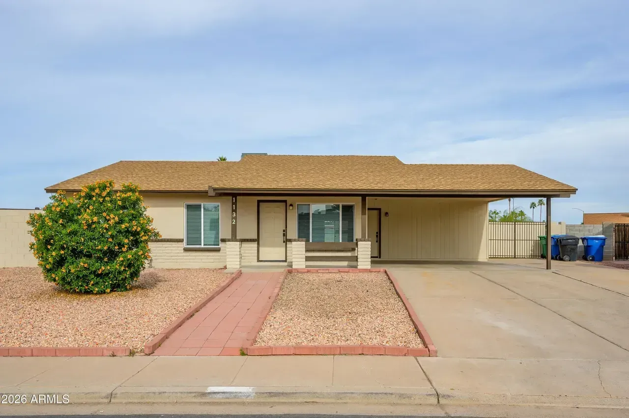Tan single-story house with a carport, tan roof, and brick walkway. Landscaped with gravel and a bush.