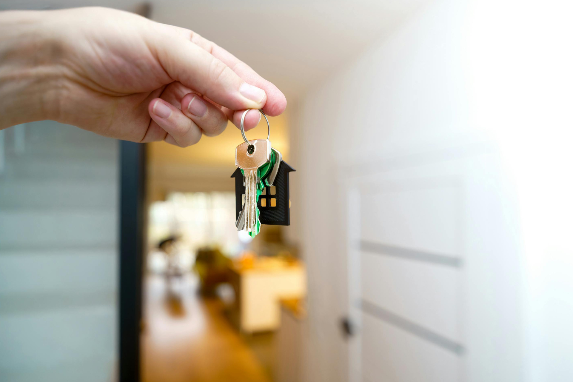 Hand holding keys with house-shaped keychain in front of a brightened interior doorway.