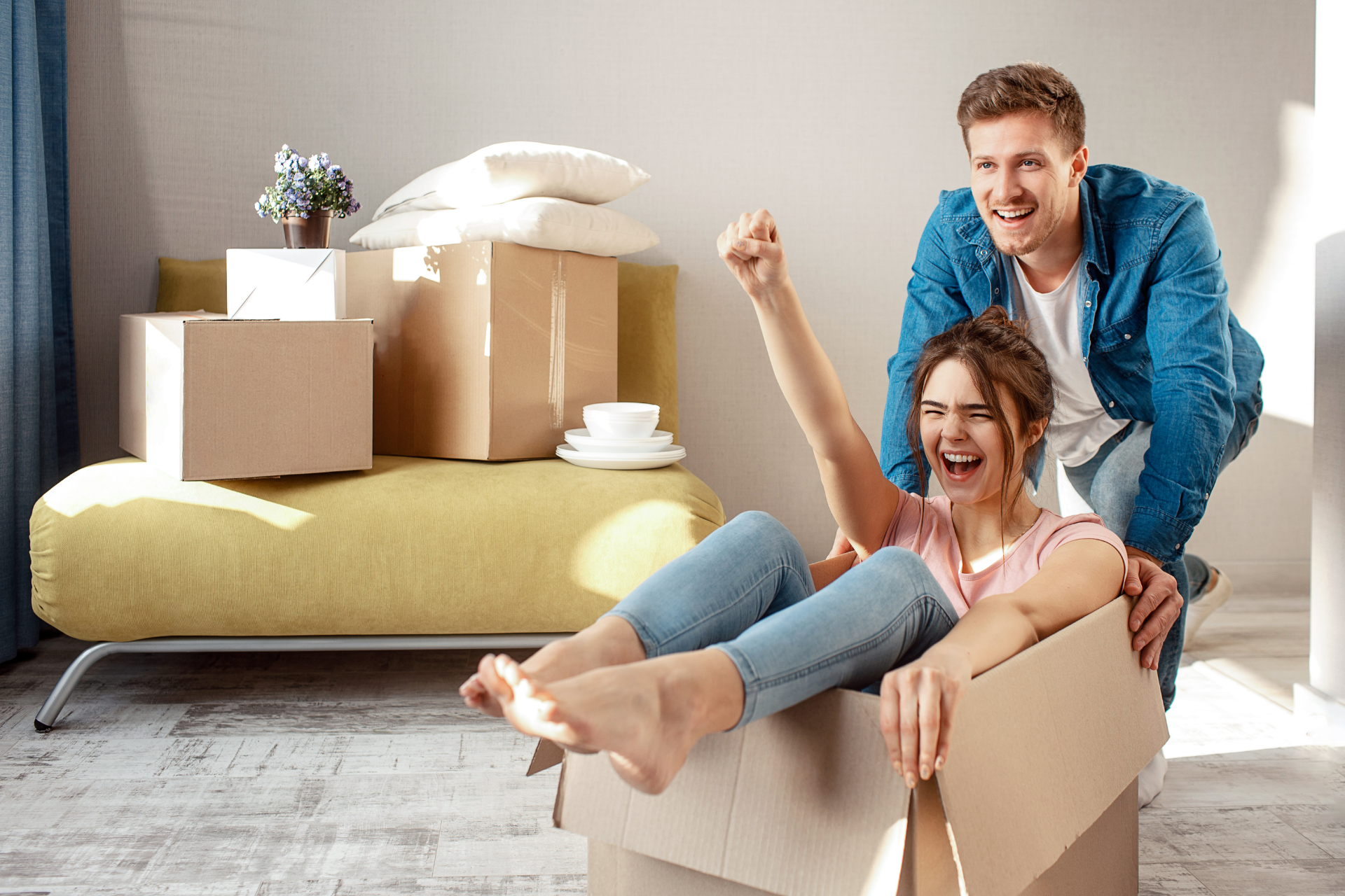 Couple celebrating in new home, woman in cardboard box cheering, man pushing, boxes, sofa.
