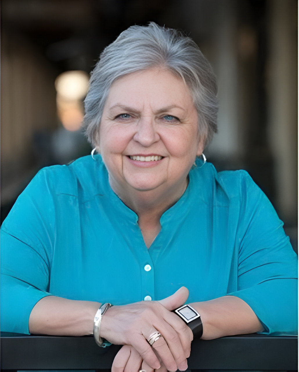 Woman with gray hair smiles, wearing a turquoise shirt, leaning on a railing.
