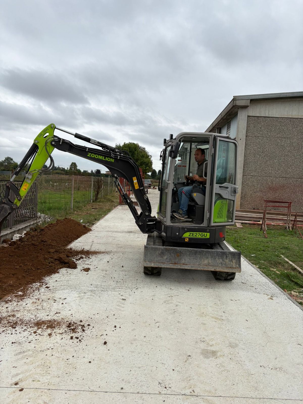 Una persona che aziona un escavatore verde e nero su una superficie di cemento, scavando terra. Cielo nuvoloso.