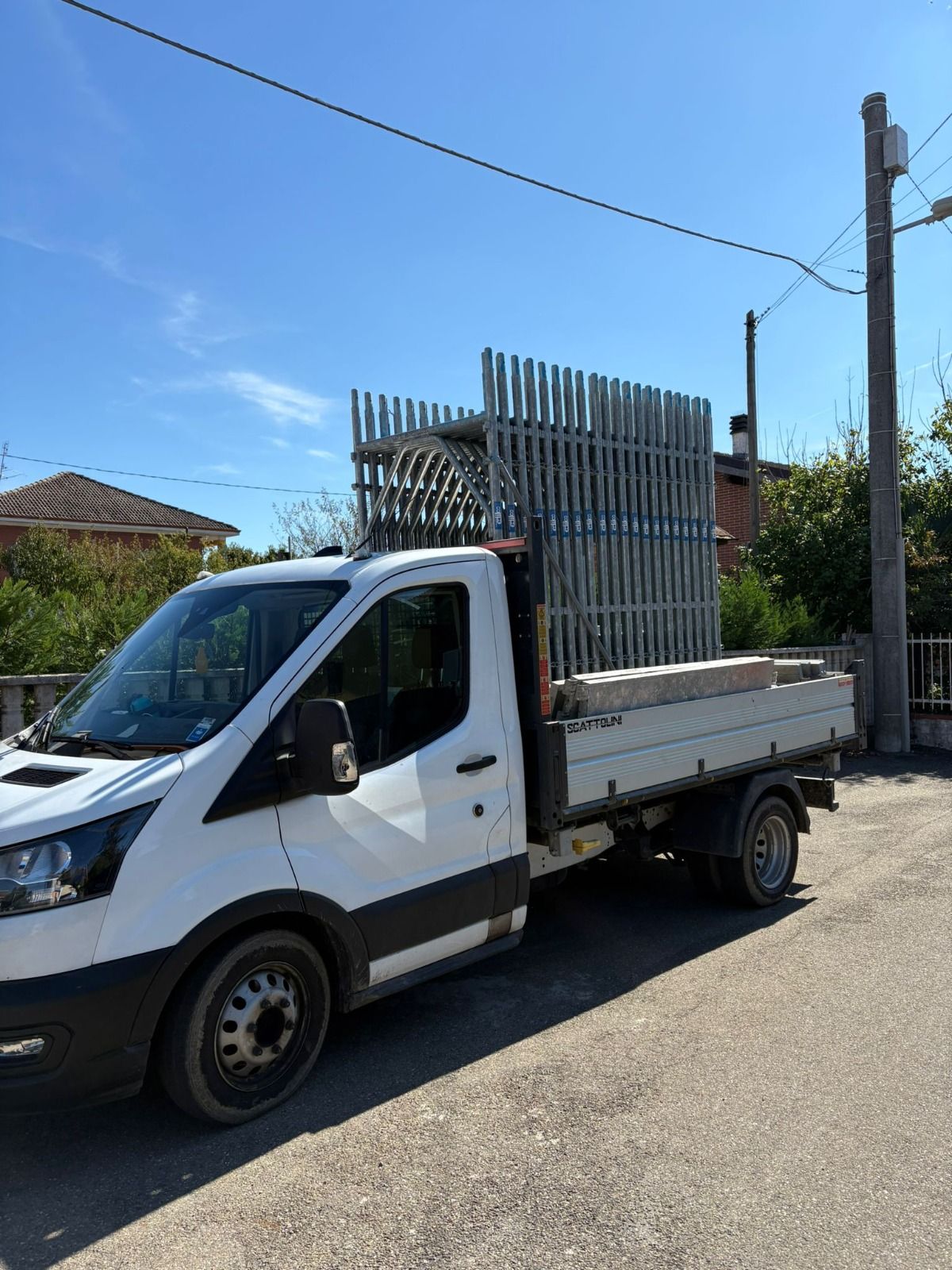 Camion Ford Transit bianco con pianale ribassato che trasporta una struttura metallica su una strada soleggiata.