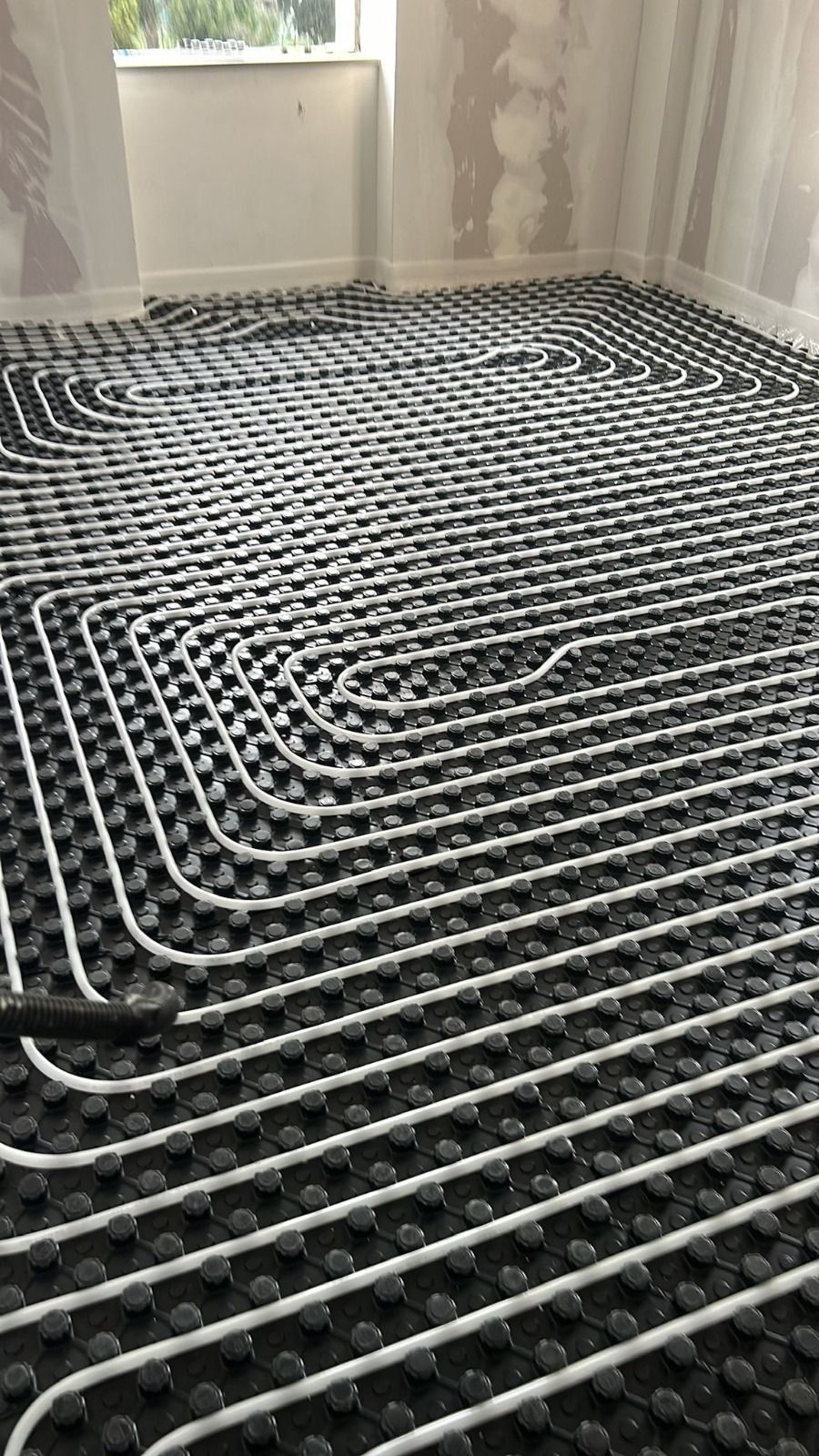 In una stanza è installato un sistema di riscaldamento a pavimento nero, con tubi bianchi disposti a griglia.