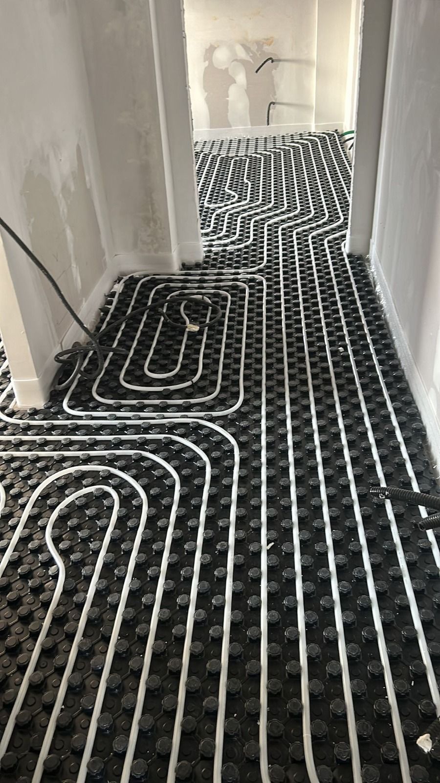 Sistema di riscaldamento a pavimento nero installato in un corridoio con pareti e porte bianche.