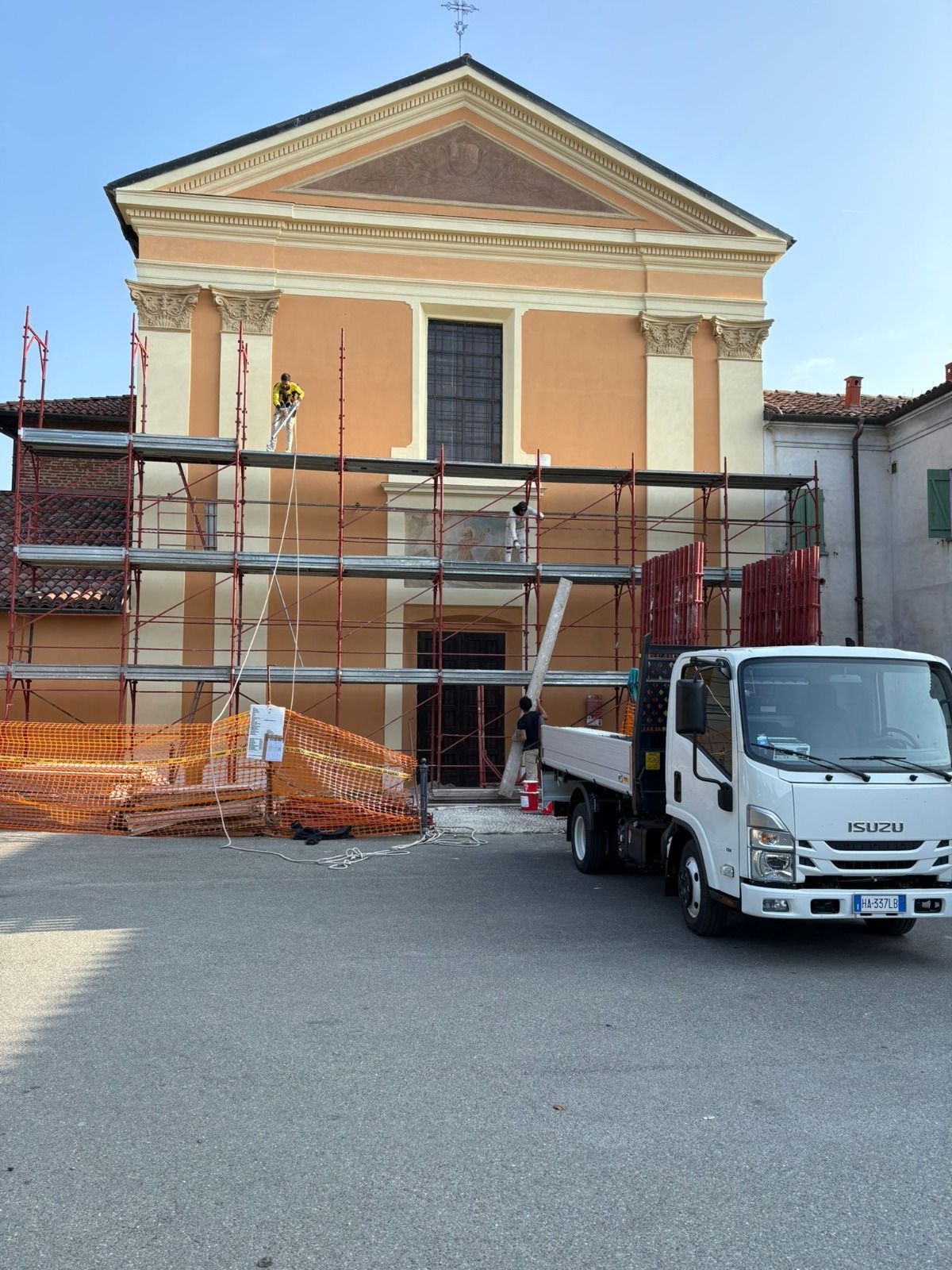 Una chiesa arancione chiaro in fase di ristrutturazione con impalcature. Un camion bianco è parcheggiato di fronte.