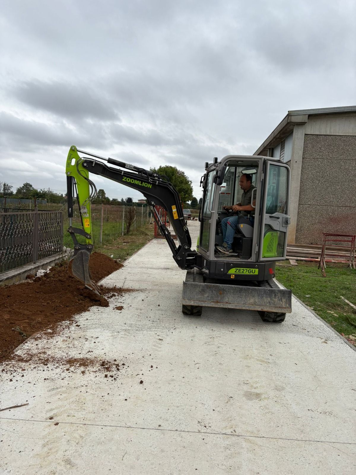 Un piccolo escavatore scava il terreno accanto a un sentiero di cemento; un uomo lo manovra. Cielo grigio sopra di noi.