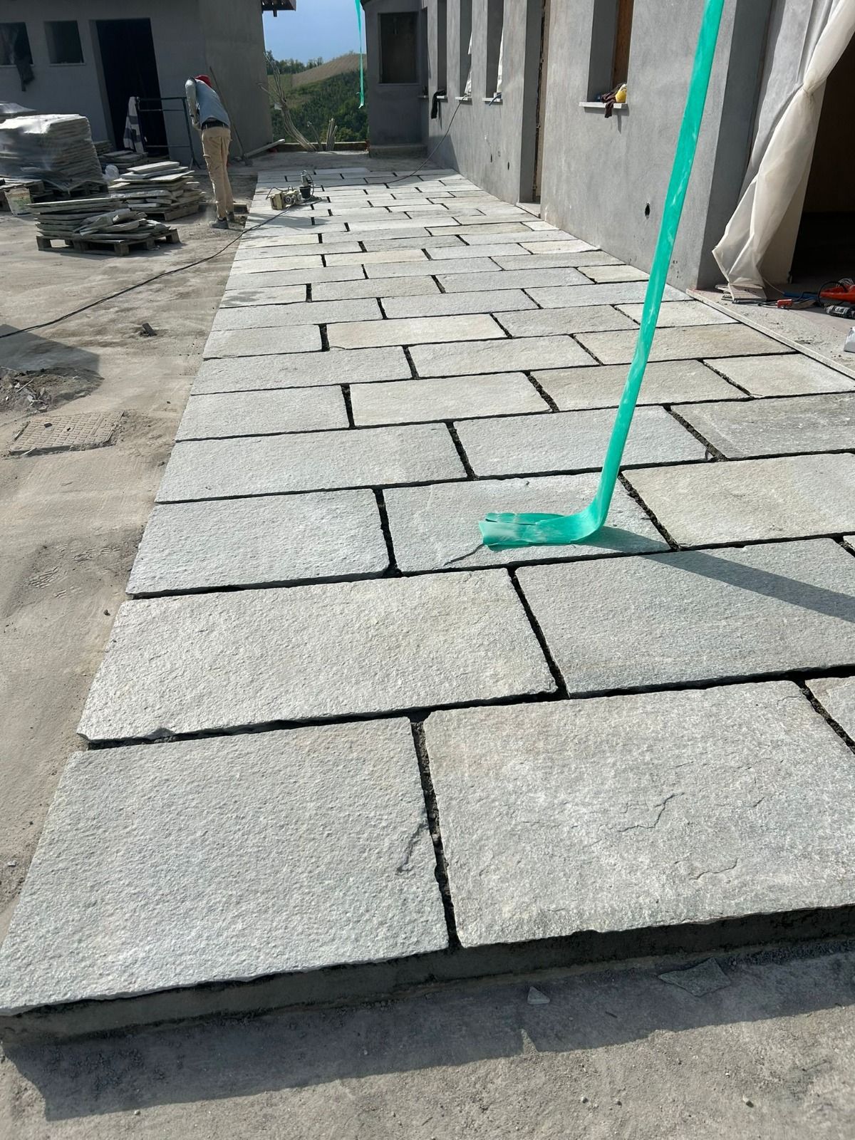 Pavimentazioni in pietra in fase di installazione su un patio in cemento. Una persona lavora sullo sfondo. Un attrezzo verde è appoggiato sulle pavimentazioni.