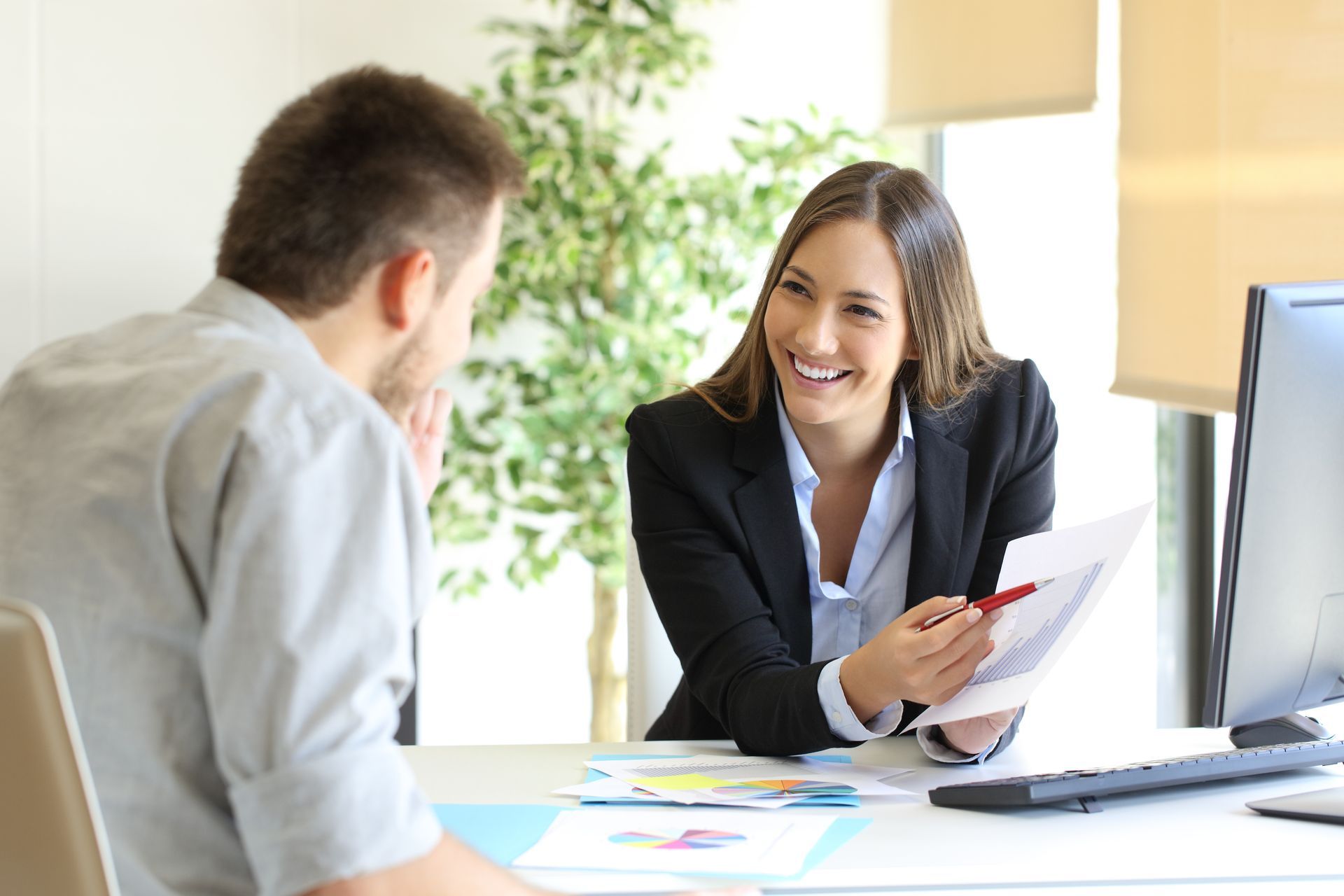 A professional woman smiles as she presents a document to a seated man across a desk in a brightly lit office.