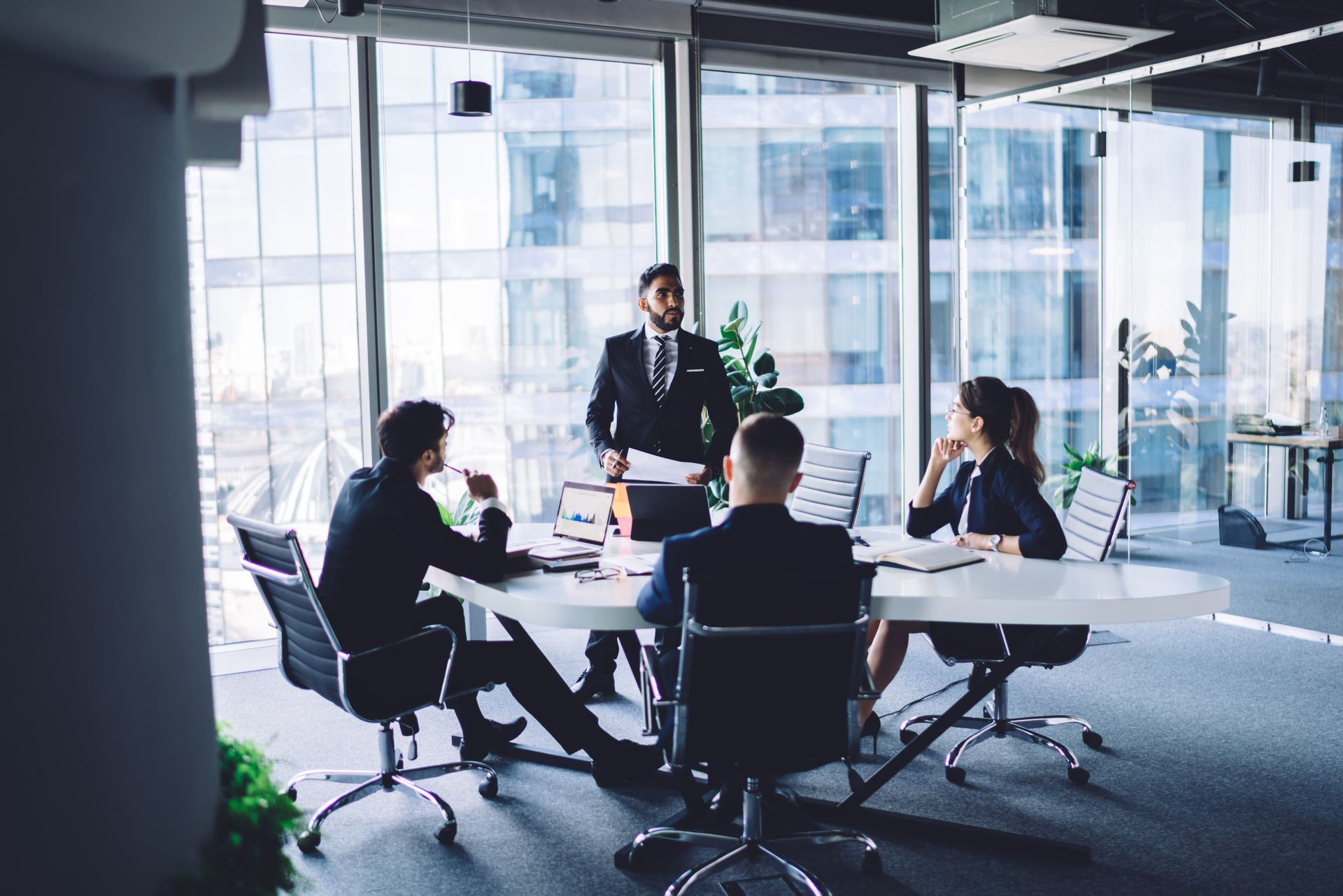 Four professionals in business attire hold a meeting around a white oval table in a modern, glass-walled office.