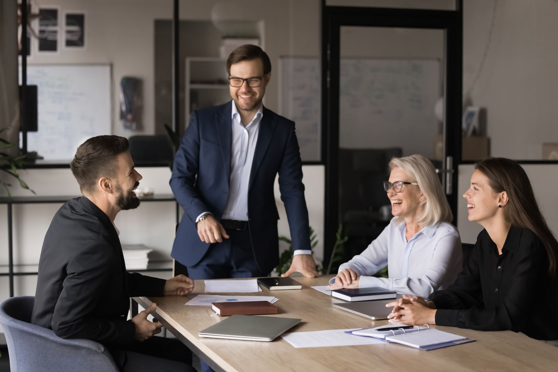 Four colleagues in professional business attire laughing together during a meeting at a wooden conference table.